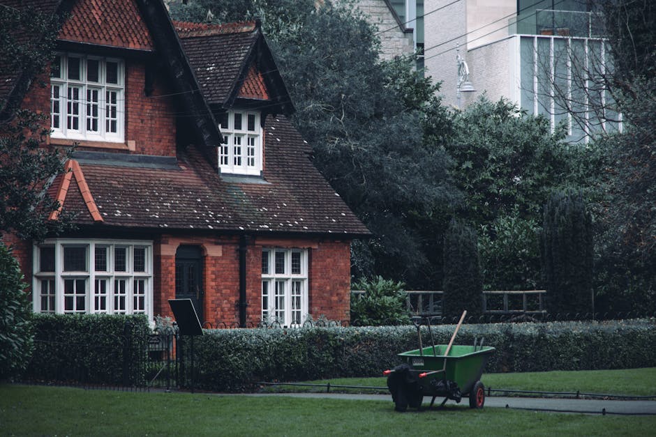 A red brick house with white-framed, multi-pane windows and a dark shingled roof surrounded by a well-maintained garden. The garden features a neatly trimmed hedge bordering a lush green lawn, with a 